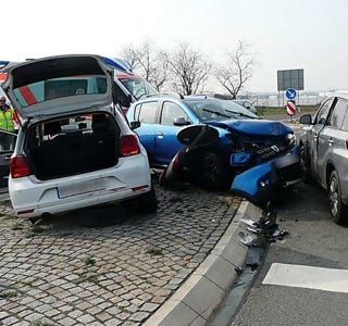 Mehrere Autos sind in einen Unfall verwickelt, mit sichtbaren Schäden an den Fahrzeugen. Ein Auto hat den Kofferraum offen. Personen in Warnwesten sind im Hintergrund zu sehen.